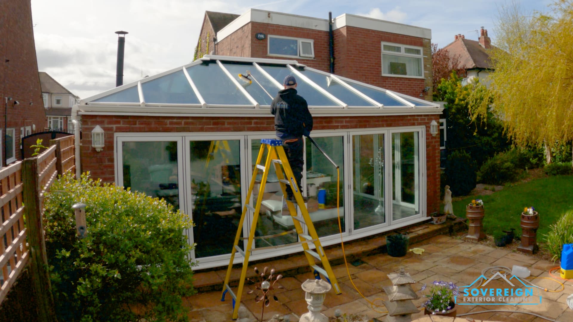 Sovereign cleaning a glass conservatory roof from a step ladder with a brush and hose